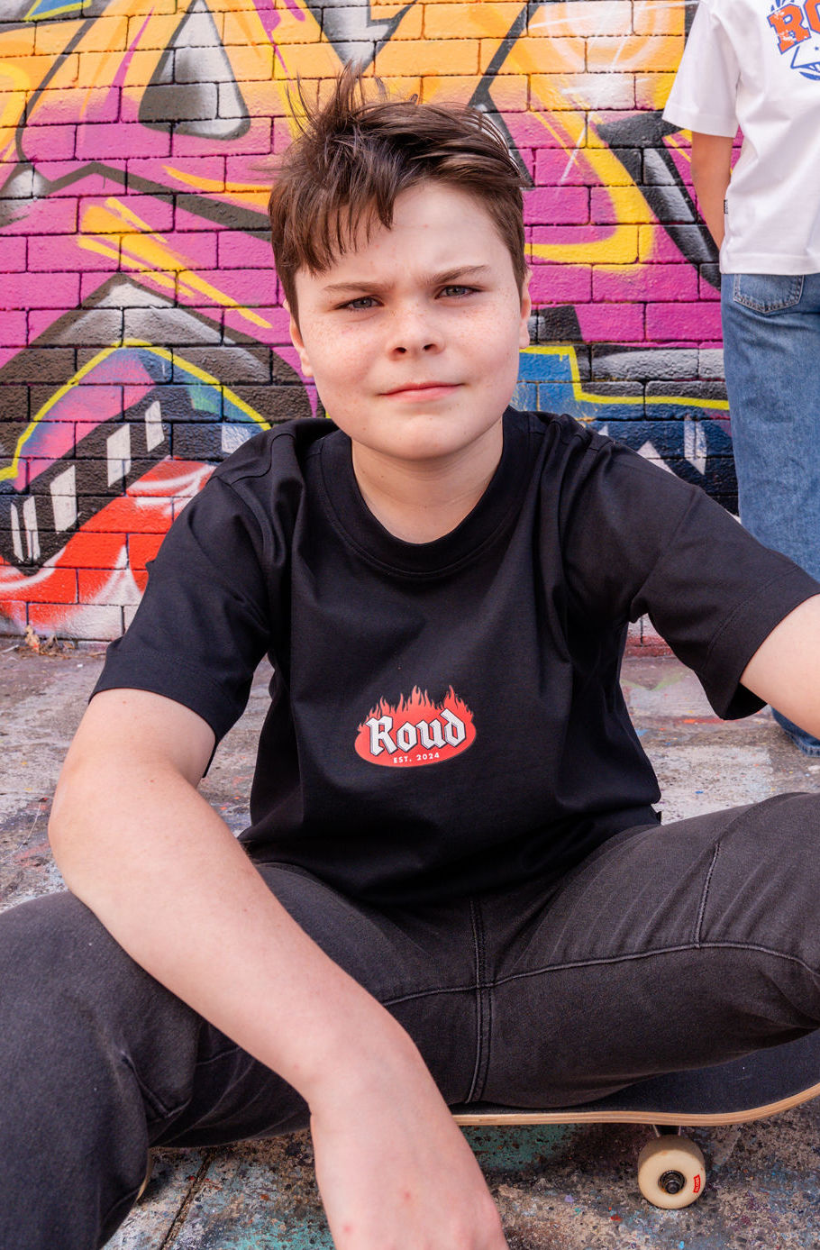 Youngman wearing a black t-shirt with a logo sitting on a skateboard against a graffiti-covered wall.